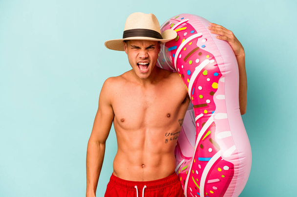 Young caucasian man holding an inflatable donut isolated on blue background screaming very angry and aggressive. - Foto, imagen