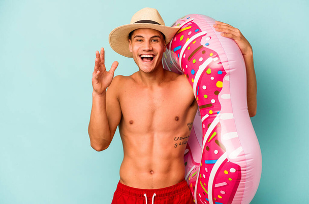 Young caucasian man holding an inflatable donut isolated on blue background receiving a pleasant surprise, excited and raising hands. - Zdjęcie, obraz