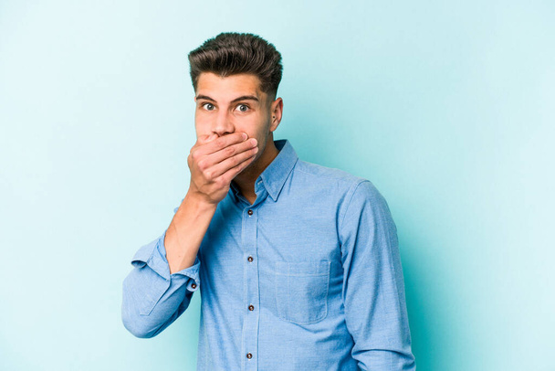 Молодой кавказский мужчина изолирован на синем фоне, прикрывая рот руками, выглядящими обеспокоенными.. - Фото, изображение