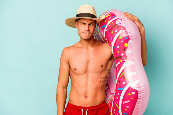 Young caucasian man holding an inflatable donut isolated on blue background confused, feels doubtful and unsure. - Foto, imagen