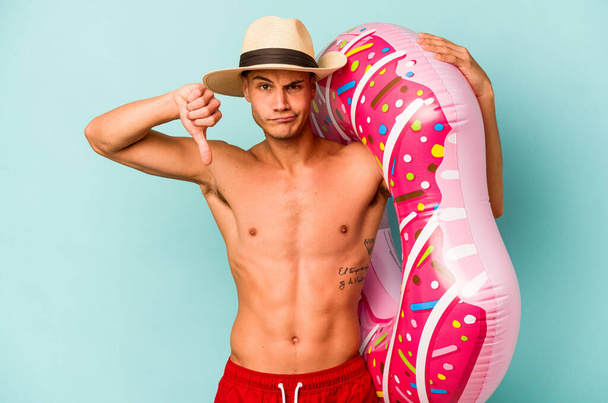 Young caucasian man holding an inflatable donut isolated on blue background showing a dislike gesture, thumbs down. Disagreement concept. - Zdjęcie, obraz