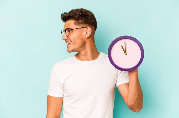 Young caucasian man holding a clock isolated on blue background looks aside smiling, cheerful and pleasant. - Foto, imagen