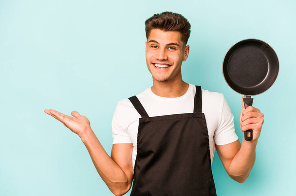 Young caucasian cooker holding frying pan isolated on blue background showing a copy space on a palm and holding another hand on waist. - Foto, imagen