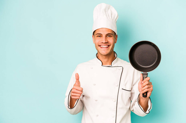 Joven chef caucásico sosteniendo la sartén voladora aislada sobre fondo azul sonriendo y levantando el pulgar hacia arriba - Foto, imagen
