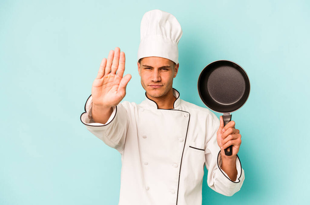 Joven chef caucásico sosteniendo la sartén voladora aislada sobre fondo azul de pie con la mano extendida mostrando señal de stop, lo que le impide. - Foto, imagen