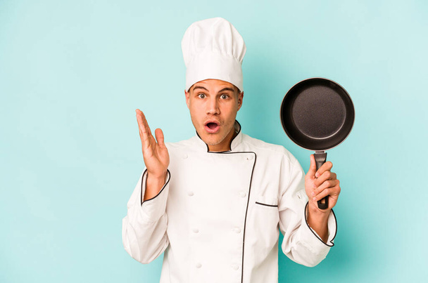 Young caucasian chef man holding flying pan isolated on blue background surprised and shocked. - Foto, immagini