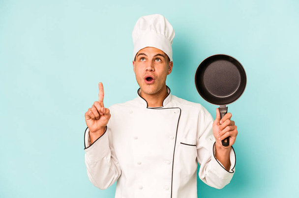 Young caucasian chef man holding flying pan isolated on blue background pointing upside with opened mouth. - Fotoğraf, Görsel