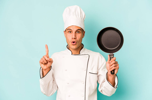 Young caucasian chef man holding flying pan isolated on blue background having some great idea, concept of creativity. - 写真・画像