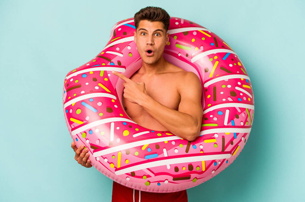 Young caucasian man holding an inflatable donut isolated on blue background pointing to the side - Foto, Bild