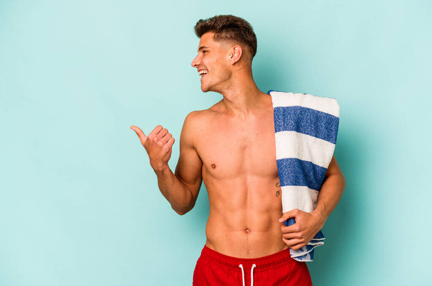 Young caucasian man holding beach towel isolated on blue background points with thumb finger away, laughing and carefree. - Foto, Imagen