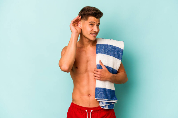 Young caucasian man holding beach towel isolated on blue background trying to listening a gossip. - Foto, Imagen