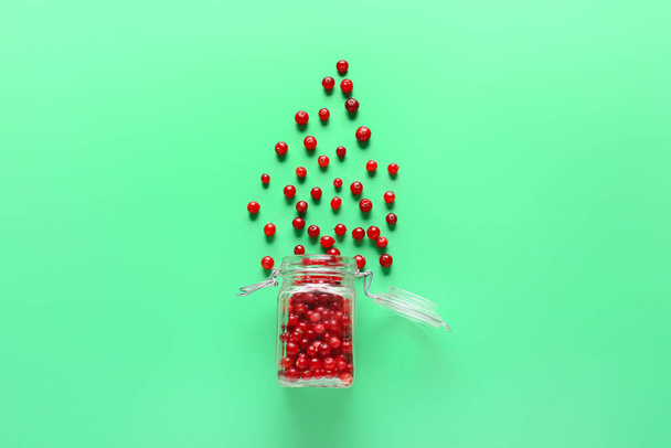 Glass jar with scattered lingonberries on green background - Foto, afbeelding