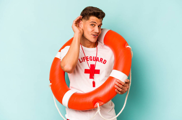 Young caucasian lifeguard isolated on blue background trying to listening a gossip. - Φωτογραφία, εικόνα