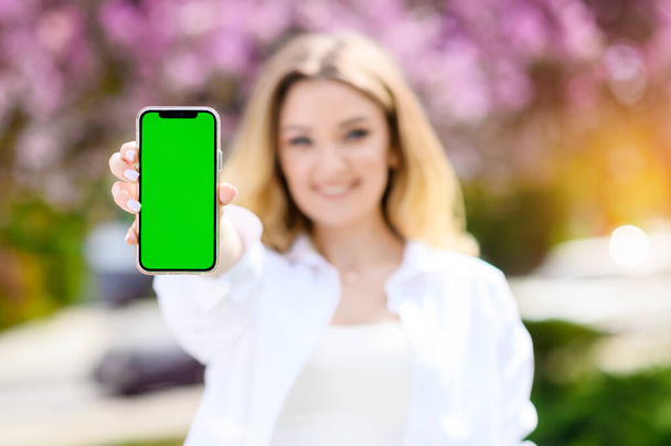 Beautiful blonde hair girl in a white shirt holding a phone with a green screen - Fotoğraf, Görsel