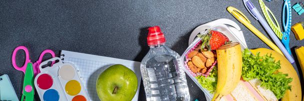 Здорова шкільна обідня коробка: бутерброд, овочі, фрукти, горіхи та йогурт з шкільними дитячими приладдям, аксесуарами та рюкзаками на чорному фоні плоскою копією простору. Повернутися до концепції школи
 - Фото, зображення