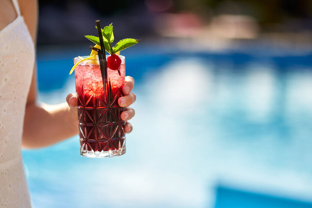Close-up of woman hand cocktail glass decorated with mint, cherry and lemon. Female holding a drink near swimming pool with blue water. Girl chilling with beverage in tropical sun. Vacation concept - Foto, Imagem