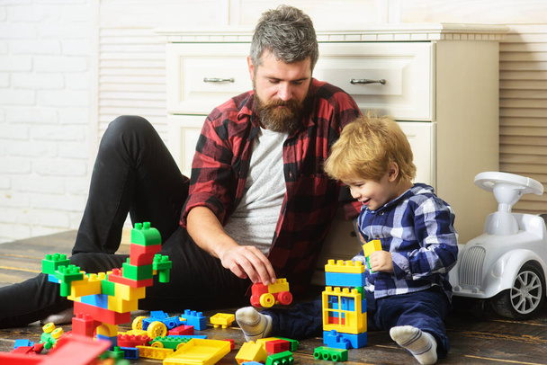 Father takes part in game with son playing on floor holding toy cars. Father and son create toys from bricks. Dad and kid build of plastic blocks. Generations family. - 写真・画像