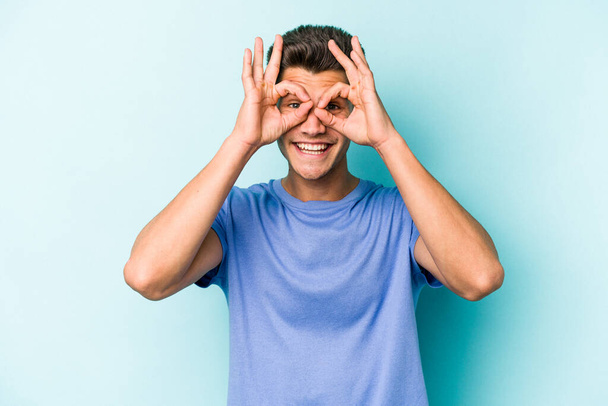 Young caucasian man isolated on blue background excited keeping ok gesture on eye. - Fotó, kép