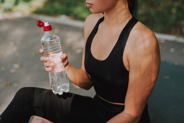 Young woman in black sportswear stretching and doing sport in park. Attractive fit girl drinking water on a break. Healthy lifestyle concept. - Fotografie, Obrázek