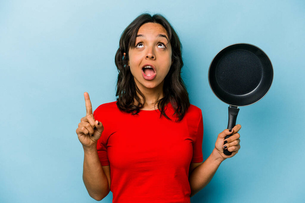 Young hispanic woman holding a frying pan isolated on blue background pointing upside with opened mouth. - Foto, immagini