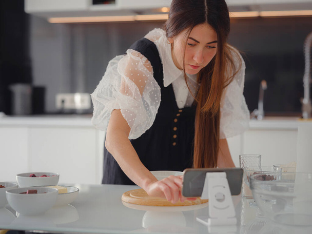 Young woman looks at a recipe for cooking a pie in a smartphone. food tutorial or cooking courses online. Baking dessert concept. - Foto, Imagen
