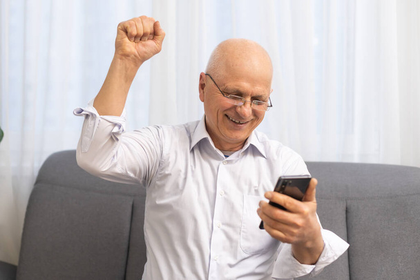 Mutlu büyükbaba akıllı telefonu tutuyor, gülüyor, sevinç çığlıkları atıyor, iyi haberler alıyor, ekranda kısa mesaj okuyor, kazanan el hareketi yapıyor. Yaşlı adamın ödülü kazanmasına şaşırdım. - Fotoğraf, Görsel