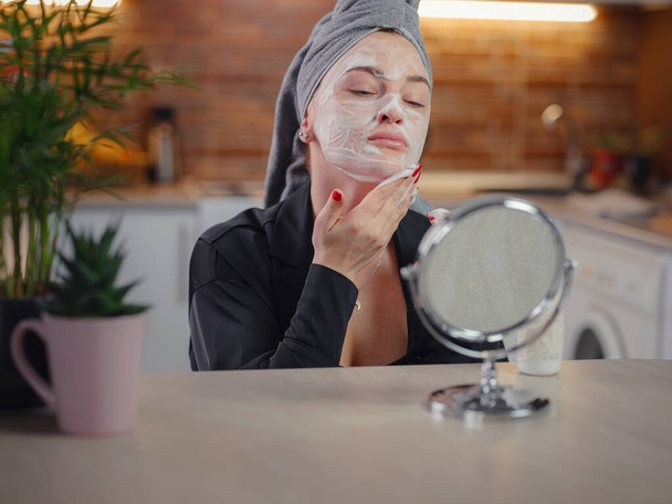 Enjoying flawless skin . Peaceful lady being satisfied with condition of her skin during procedures in kitchen. Beautiful caucasian woman is cleaning face with Cleansing foam looking in mirror. - Foto, Imagem