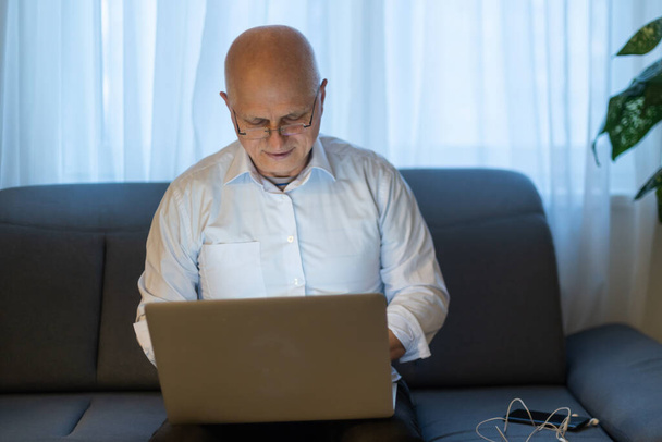 Літній лисий чоловік у білій сорочці і чорні штани виправляють окуляри і працюють на ноутбуці, сидячи у вітальні
. - Фото, зображення