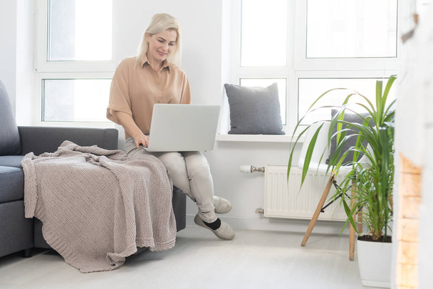Uśmiechnięta szczęśliwa kobieta siedząca na kanapie i korzystająca z laptopa. Pojęcie pracy na odległość lub nauki podczas kwarantanny - Zdjęcie, obraz