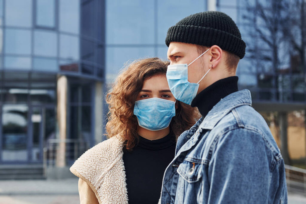 Couple in protective masks have a walk outdoors in the city near business building at quarantine time. Conception of coronavirus. - Foto, imagen