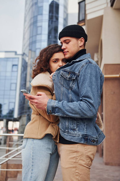 Using phone. Cheerful couple in casual warm clothes have a walk outdoors in the city near business building. - Fotografie, Obrázek