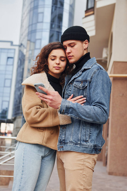 Using phone. Cheerful couple in casual warm clothes have a walk outdoors in the city near business building. - Foto, Imagen