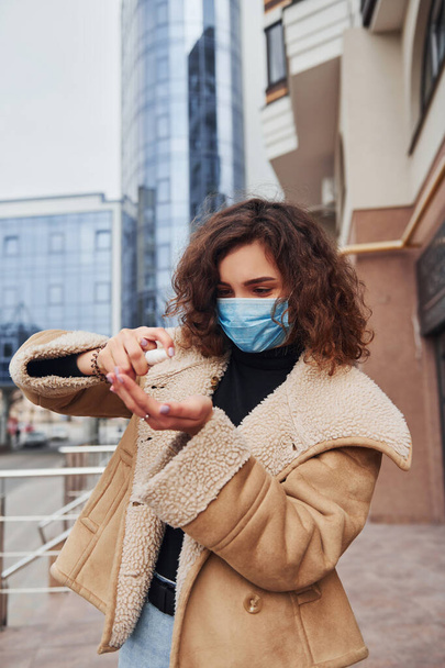 Portrait of young girl with curly hair in protective mask that using antiseptic outdoors near business building at quarantine time. Conception of coronavirus. - Foto, imagen