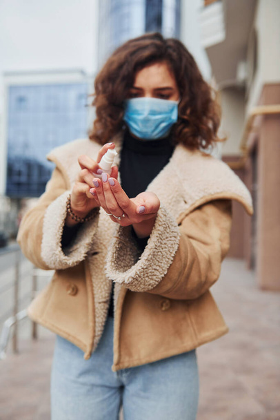 Portrait of young girl with curly hair in protective mask that using antiseptic outdoors near business building at quarantine time. Conception of coronavirus. - Фото, зображення
