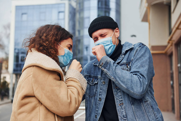 Couple in protective masks have a cough outdoors in the city near business building at quarantine time. Conception of coronavirus. - Фото, изображение