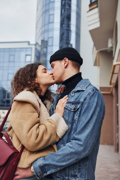 Cheerful couple in casual warm clothes kissing outdoors in the city near business building. - Foto, imagen