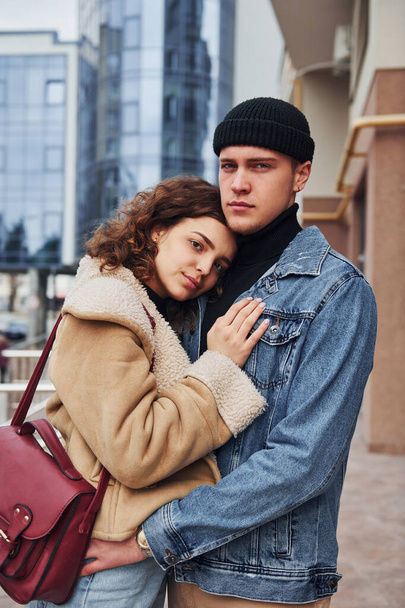 Cheerful couple in casual warm clothes embracing each other outdoors in the city near business building. - Φωτογραφία, εικόνα