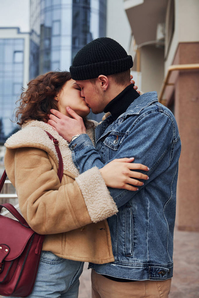 Cheerful couple in casual warm clothes kissing outdoors in the city near business building. - Foto, Imagem