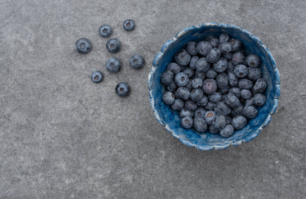 On top shot of a homemade blue clay bowl filled with fresh blueberries. There are also a few berries on the gray stone background. There is space for text on the side. - 写真・画像