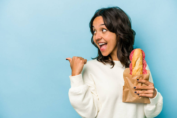 Young hispanic woman eating a sandwich isolates on blue background points with thumb finger away, laughing and carefree. - Foto, Imagen