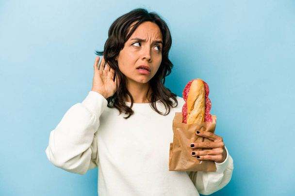jonge latino vrouw eten een sandwich isolaten op blauwe achtergrond proberen om een roddel luisteren. - Foto, afbeelding