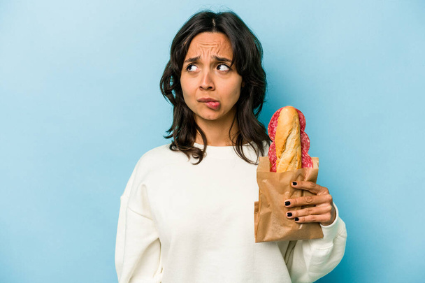 Νεαρή Ισπανίδα που τρώει σάντουιτς απομονώνεται σε μπλε φόντο μπερδεμένη, αισθάνεται αμφίβολη και αβέβαιη. - Φωτογραφία, εικόνα