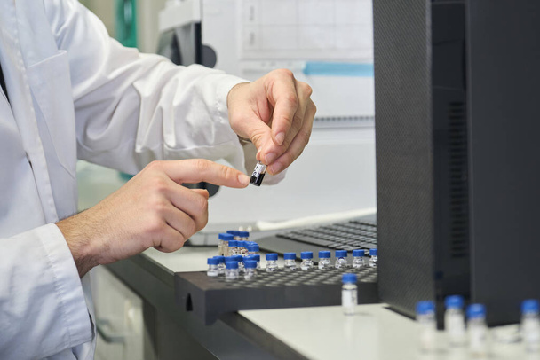 Naukowiec w fartuchu laboratoryjnym umieszcza fiolkę z próbką w autoosamplerze systemu HPLC. - Zdjęcie, obraz