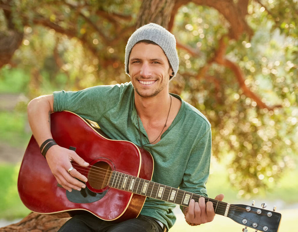 Nature inspires his music. Portrait of a handsome young man sitting outside in a park playing guitar. - Foto, immagini
