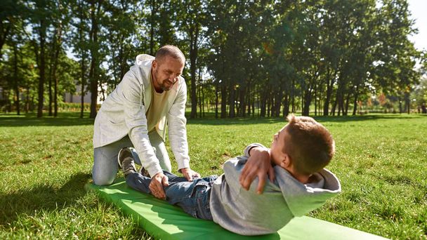 Dad help teenage son crunch on fitness mat in park - Фото, изображение