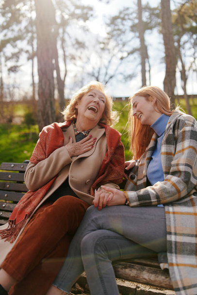 Una giovane donna rossiccia è seduta con la nonna e parla sulla panchina del parco con il freddo. - Foto, immagini