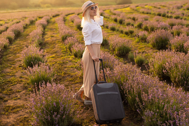 Beautiful young woman in lavender field standing with suitcase - Foto, afbeelding