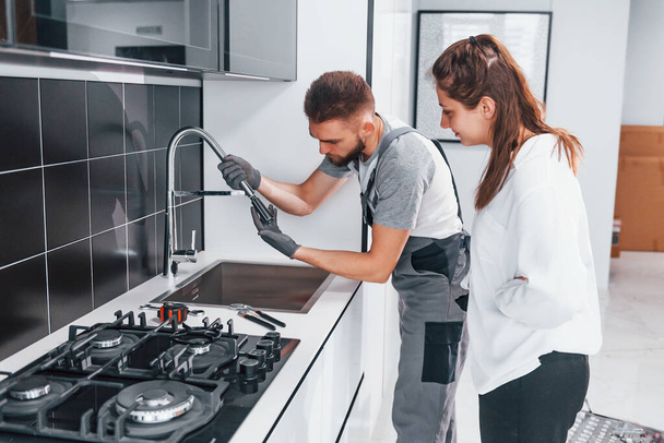 Young professional plumber in grey uniform shows results of work for housewife on the kitchen. - Φωτογραφία, εικόνα