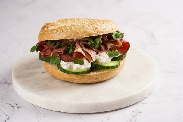 Ein Bagel-Sandwich mit Pastrami, Gurkenscheiben, Brunnenkresse-Salat und Ricotta auf Marmorgrund - Foto, Bild