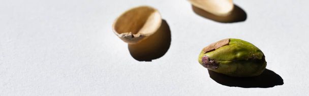 cracked green and salty pistachio near blurred nutshells on white background, banner - Фото, изображение